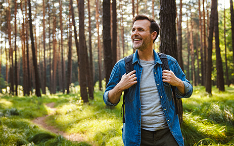 Person wearing a denim overshirt and backpack stands on a sunlit forest trail, surrounded by tall pine trees and green grass. Warm sunlight filters through the trees, illuminating the wooded landscape.