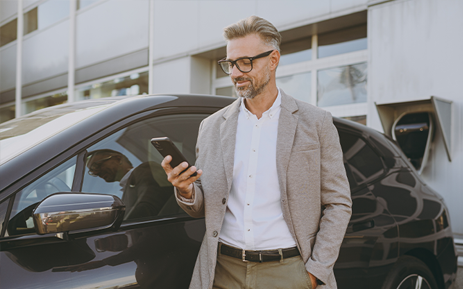 Person in business-casual clothing stands beside a black SUV outside a modern building while viewing a smartphone.