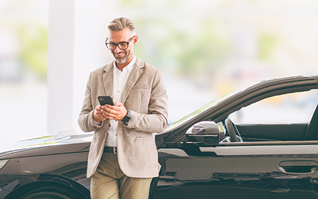 Person in business-casual clothing stands beside a black SUV outside a modern building while viewing a smartphone.