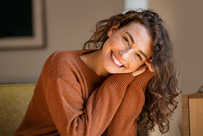 Person with curly hair wearing a brown knit sweater rests their head on folded hands in a warmly lit indoor portrait.