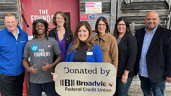 Group of seven people posing in front of The Foundry sign, holding a Donated By Broadview FCU sign