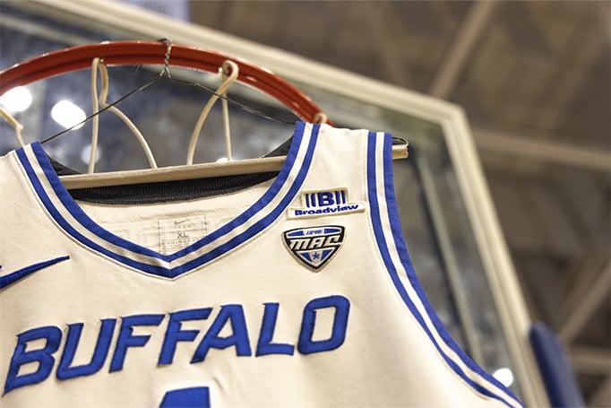 Close-up of a Buffalo basketball jersey hanging from a basketball hoop, showing the Broadview sponsor patch, MAC conference logo patch, and blue "BUFFALO" lettering on a white jersey.