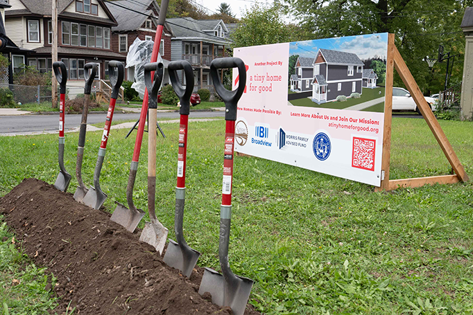 Row of ceremonial shovels lined up beside freshly turned soil at a tiny home project groundbreaking site, with a project sign displaying a rendering of the future home in the background.