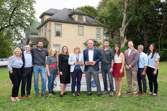Group of community partners and supporters standing in front of a vacant lot and a large house during a groundbreaking event for a tiny home project, with two people in the center holding commemorative bricks and a project sign visible in the background.