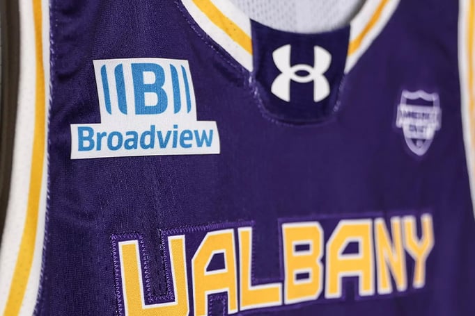 Close-up of a purple UAlbany basketball jersey featuring the Broadview logo patch, Under Armour logo, and gold-and-white "ALBANY" lettering on the front.