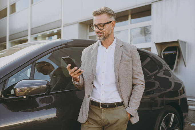 A person wearing a light gray blazer, white button-down shirt, and tan pants stands beside a black electric vehicle, looking at a smartphone. The vehicle is parked outside a modern building with large glass windows, and a charging station is visible in the background. Bright daylight reflects off the car’s glossy surface.